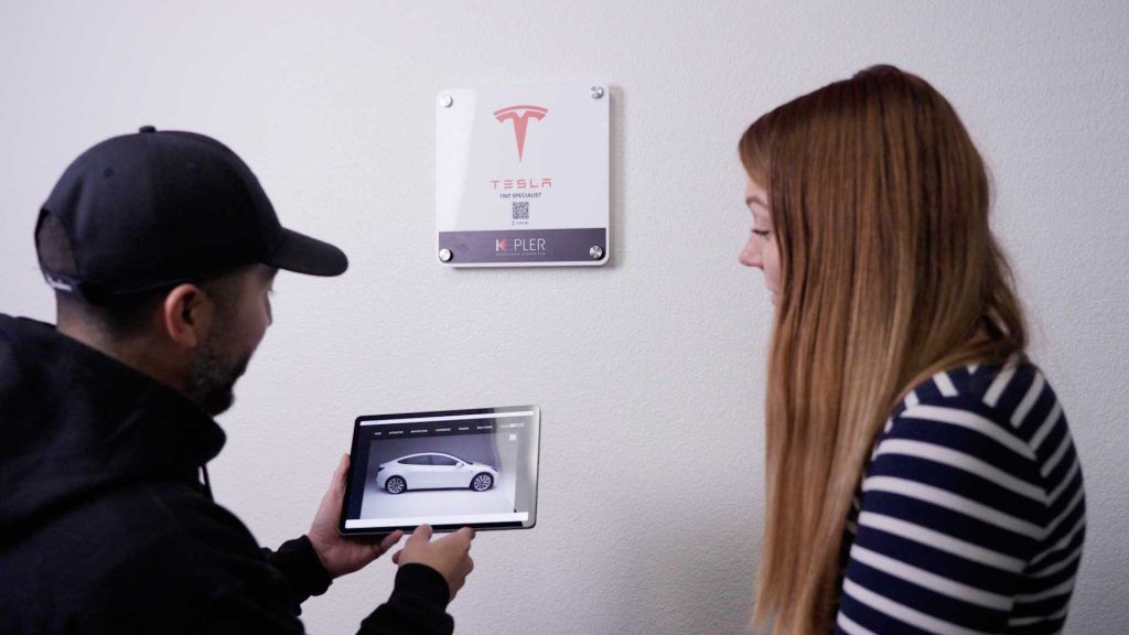 A person displays a car configuration on a tablet to another individual next to a Tesla wall sign.