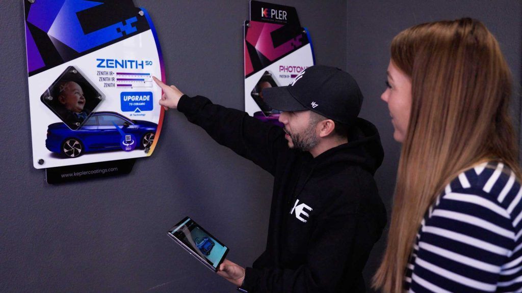 A person points to a product display on a wall while holding a tablet, with another person looking on in a room.