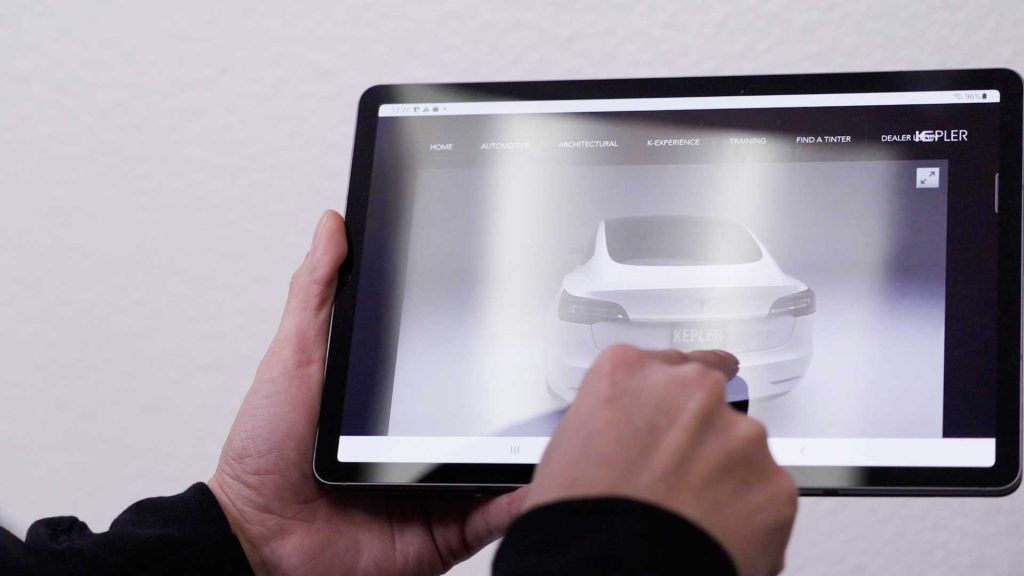 Hands holding a tablet displaying a 3D digital model of a white car, with a finger touching the screen.