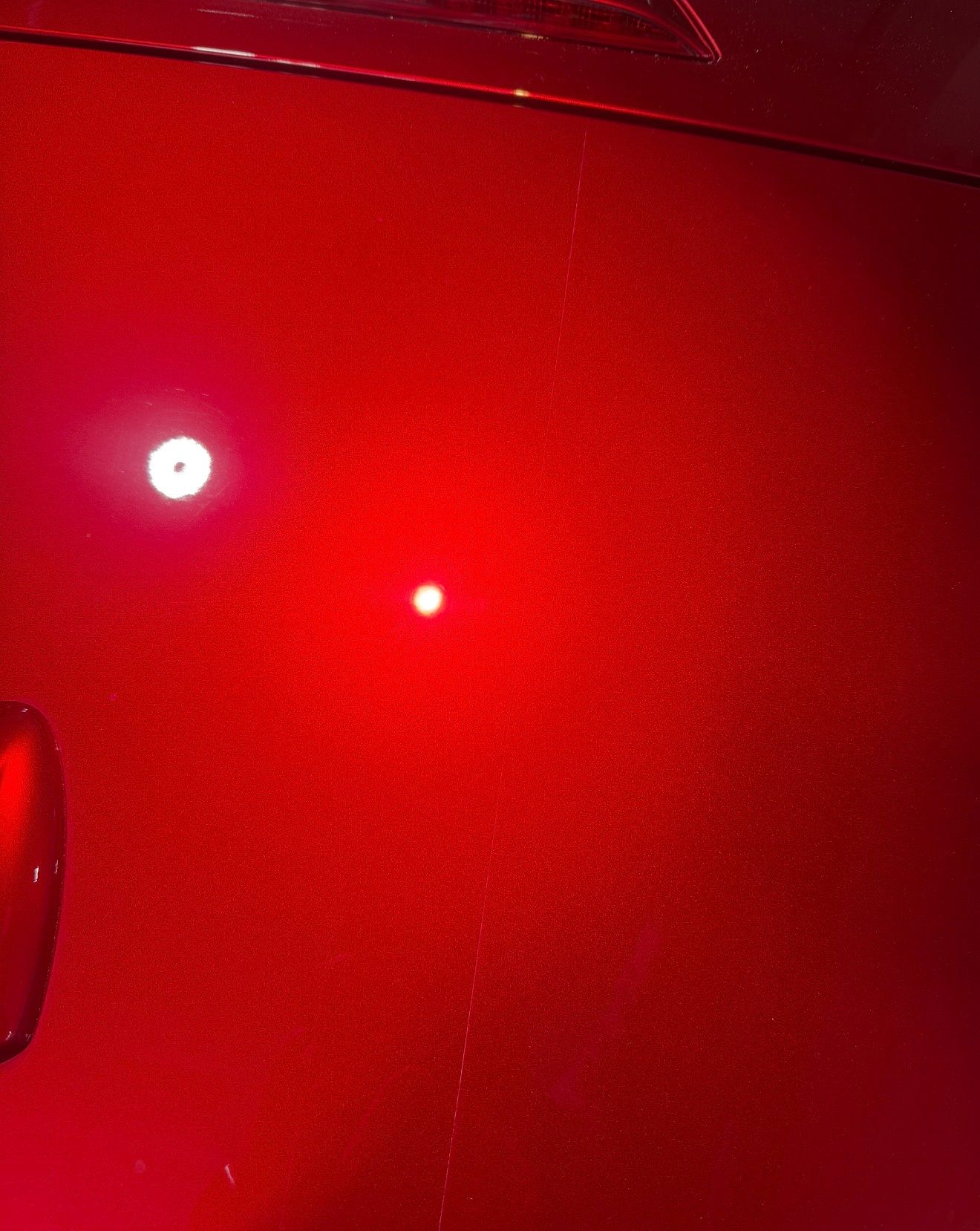 A close-up of a glossy, metallic red car surface with light reflections and a subtle vertical scratch in the center.