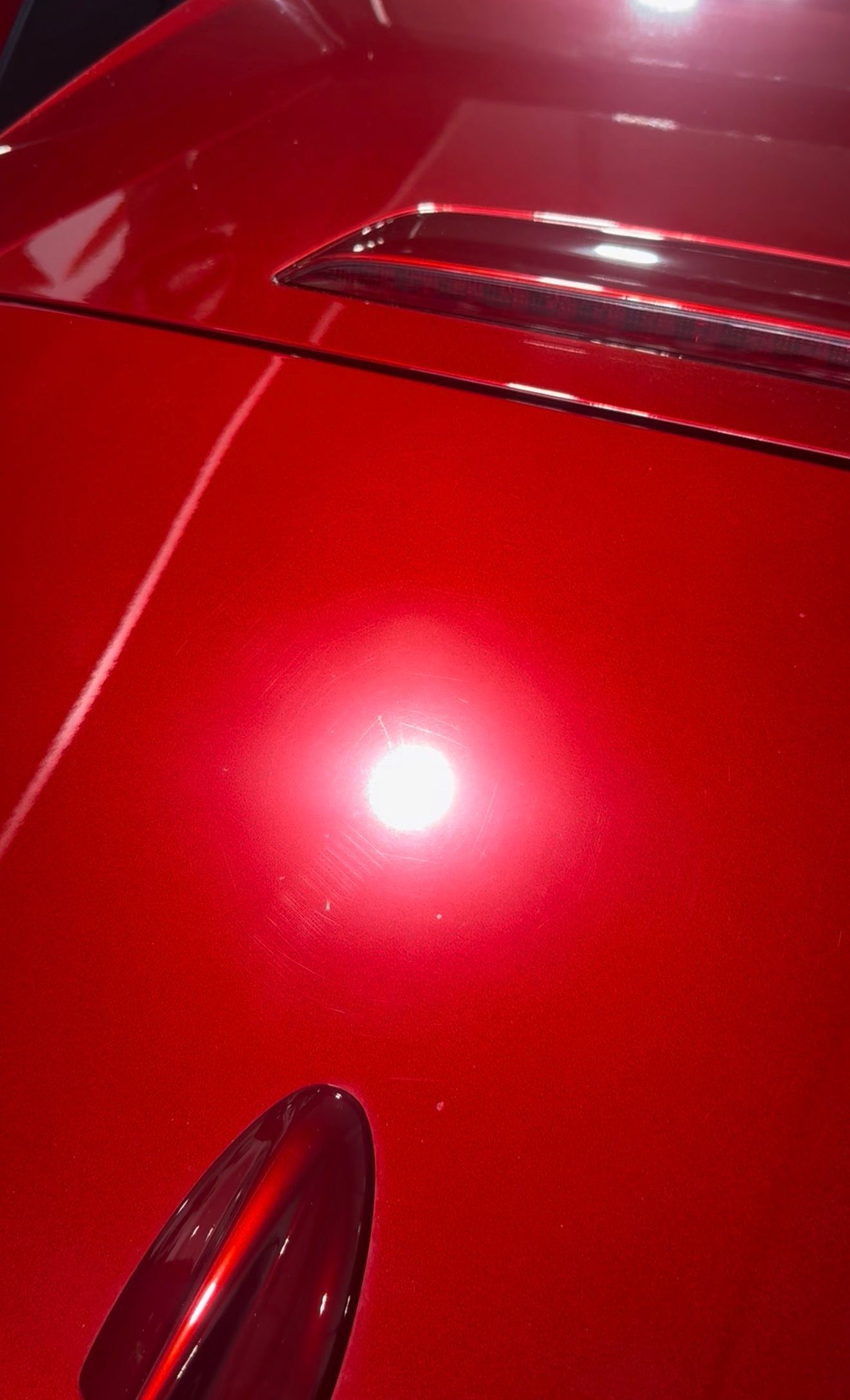 A close-up, high-angle shot of a shiny, metallic red car hood with a light reflection and a dark antenna base.