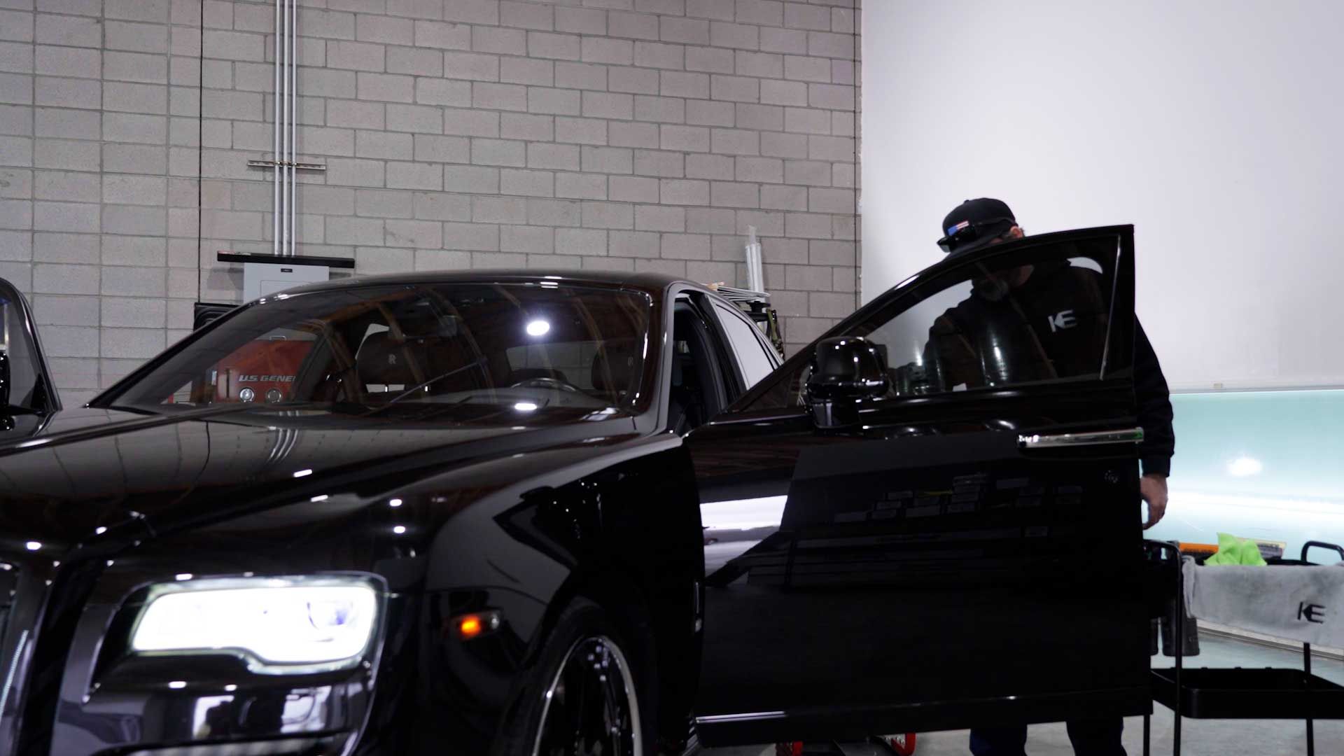 A person wearing a hat stands by the open driver’s side door of a shiny black car inside a garage with brick walls.