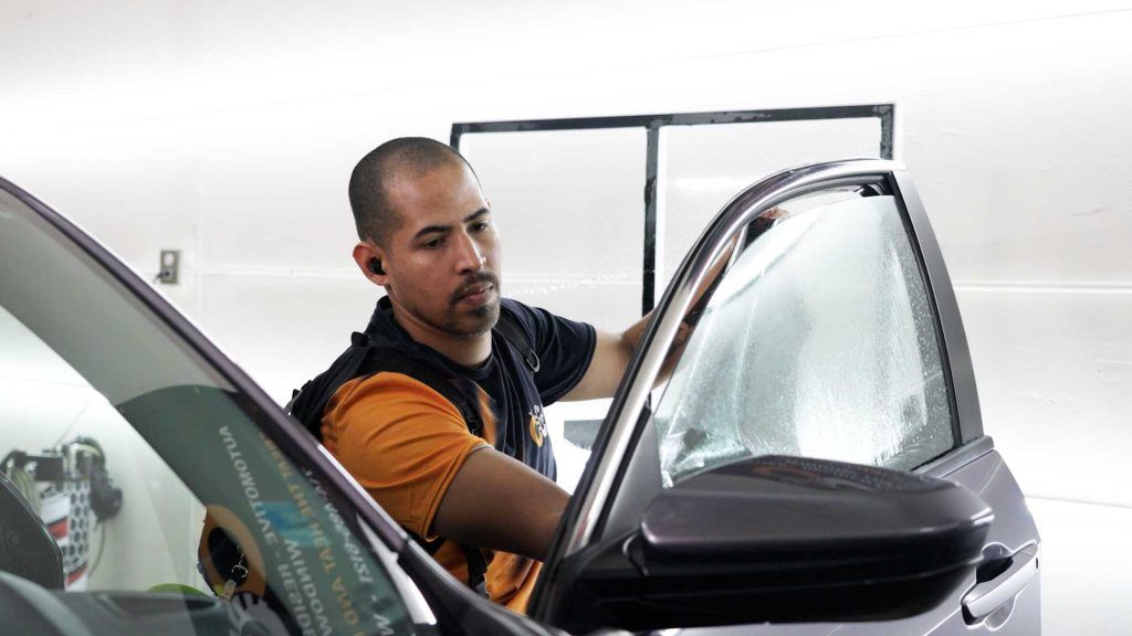 A worker in a yellow shirt applying a clear window tint film to a car door window in an indoor shop.