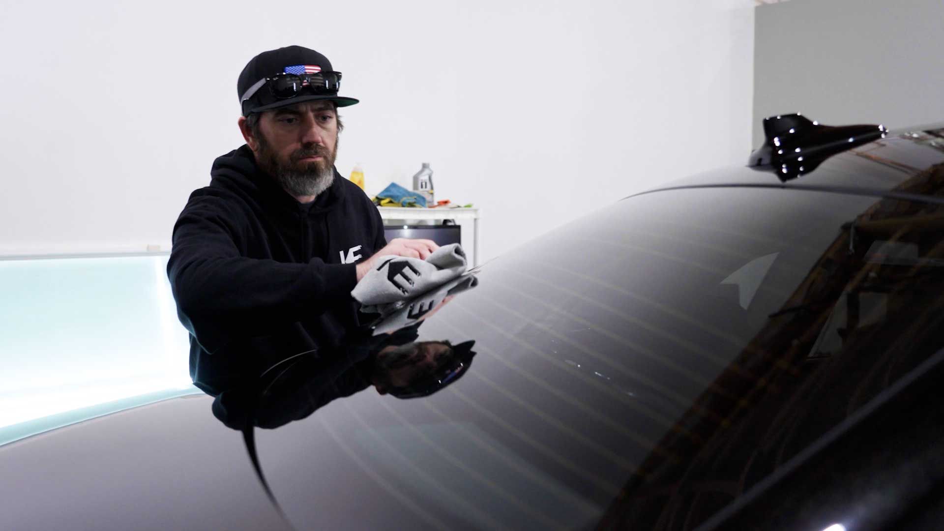 A person wearing a black hoodie and cap wipes the rear window of a black car with a white cloth in a brightly lit garage.
