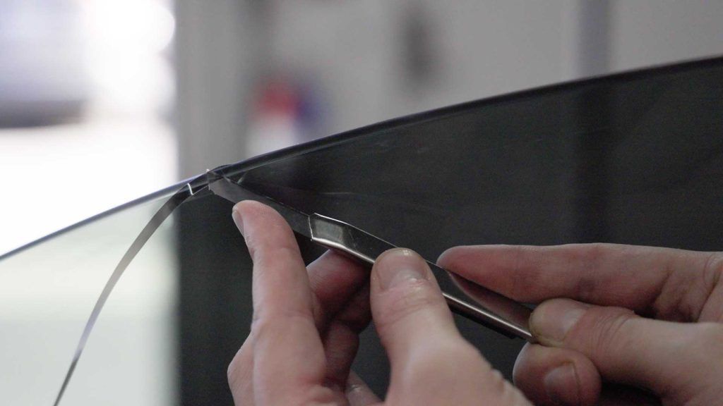 Hands use a metal tool to carefully pry or lift the edge of a thin, dark screen panel from its frame.