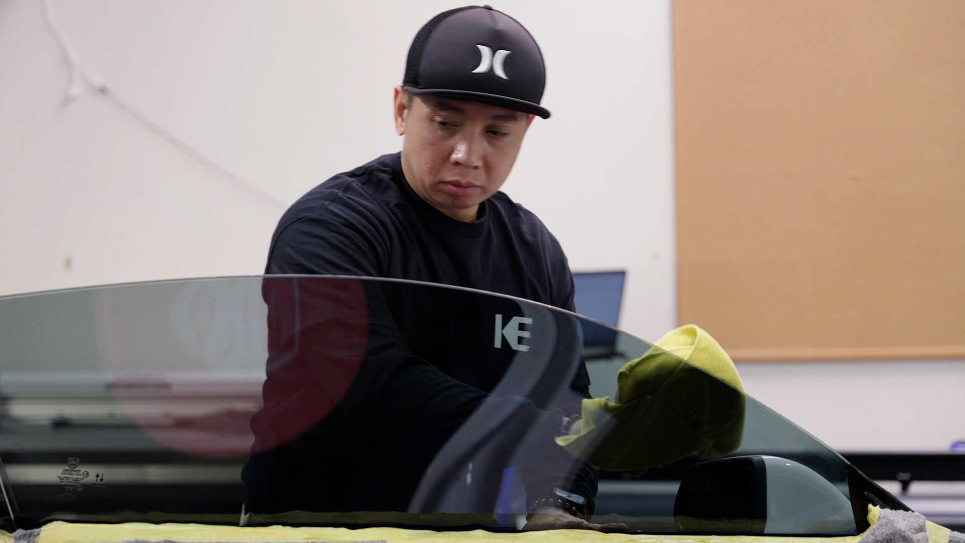 A person wearing a baseball cap carefully wipes down a car window with a cloth in a workshop setting.