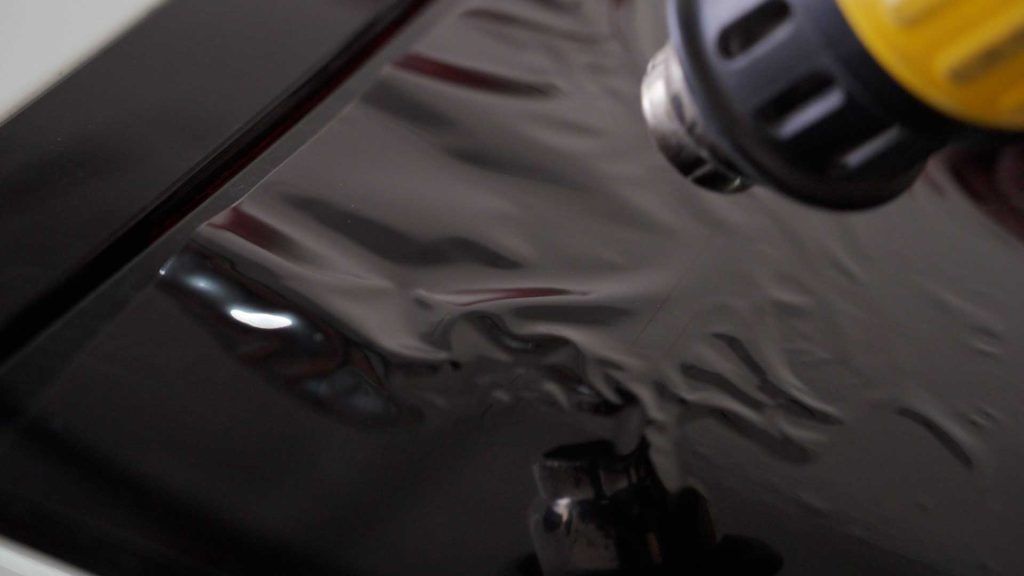A heat gun being used to smooth out black, wrinkled vinyl wrap on a car surface.
