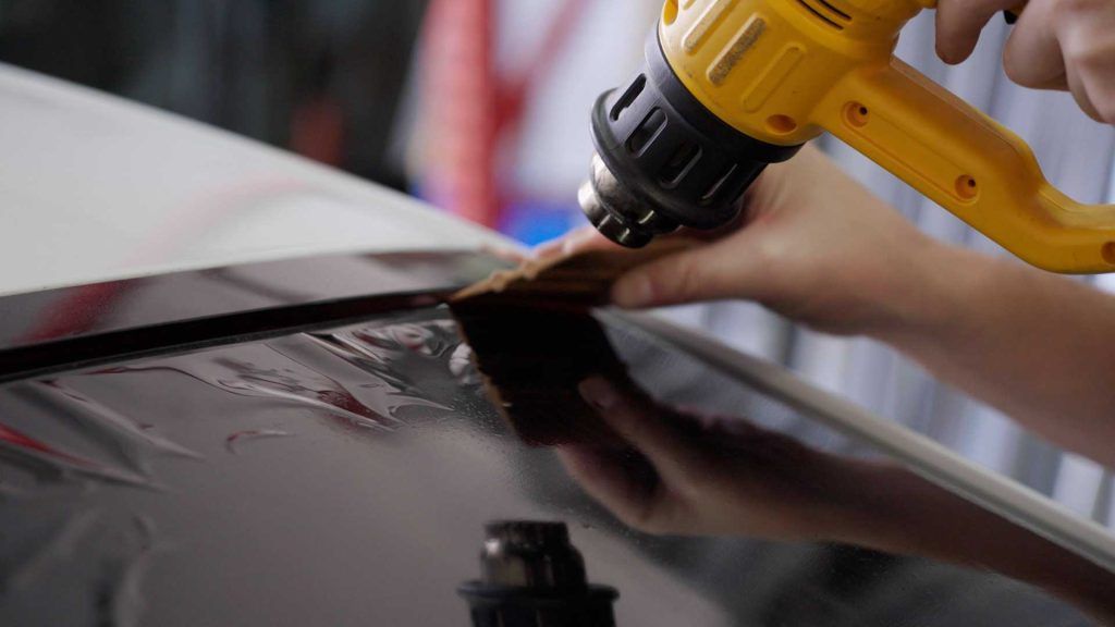 A person uses a yellow heat gun and a squeegee to apply a dark adhesive film to a car roof.