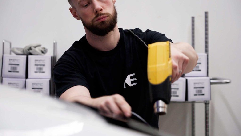 A person using a yellow heat gun to apply film to a car surface in a workshop with boxes on shelves in the background.