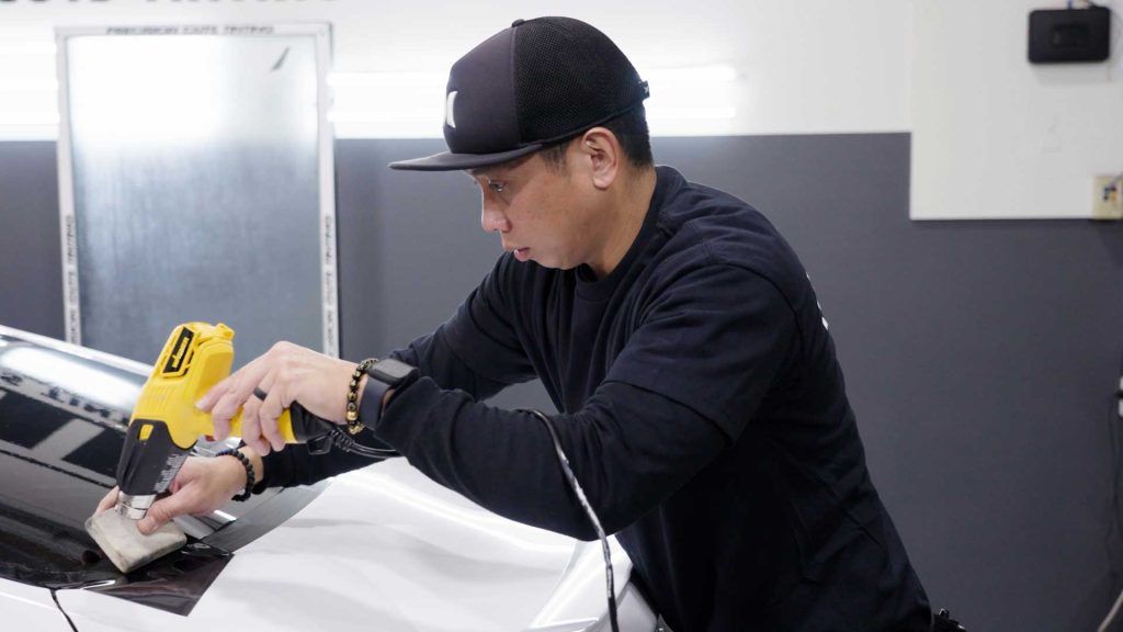 A person in a black long-sleeve shirt and cap uses a yellow heat gun to apply white vinyl wrap to a vehicle surface.