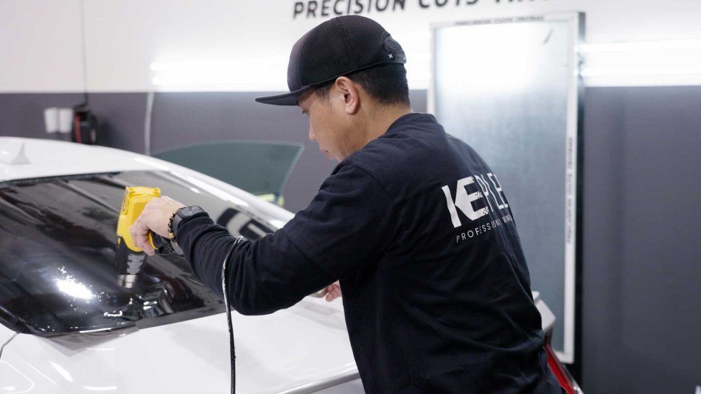 A technician uses a yellow heat gun to apply window tint to the rear window of a white car in a garage.