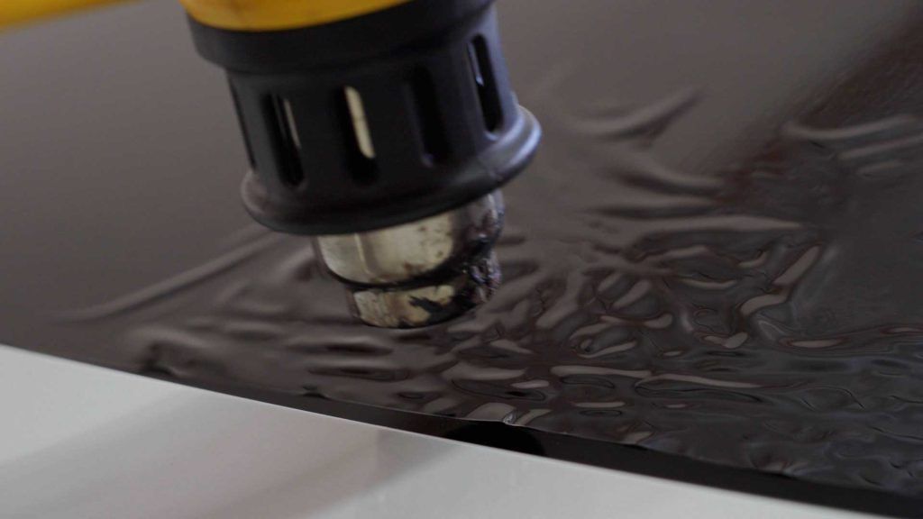 A heat gun applies heat to a black, textured vinyl wrap sheet to make it pliable for application.