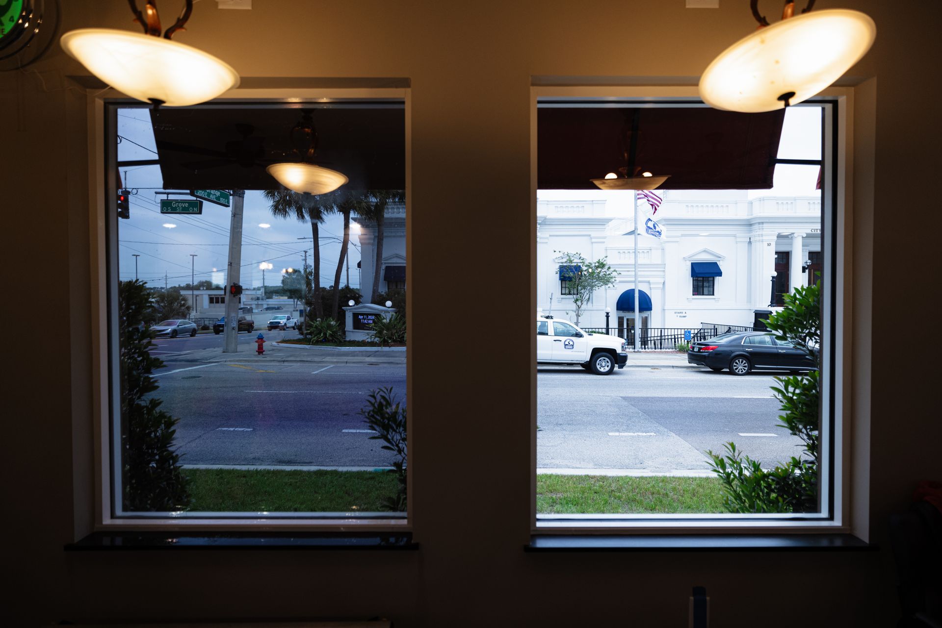 A view from indoors through two windows showing a street, a white building, and cars, illuminated by hanging light fixtures.