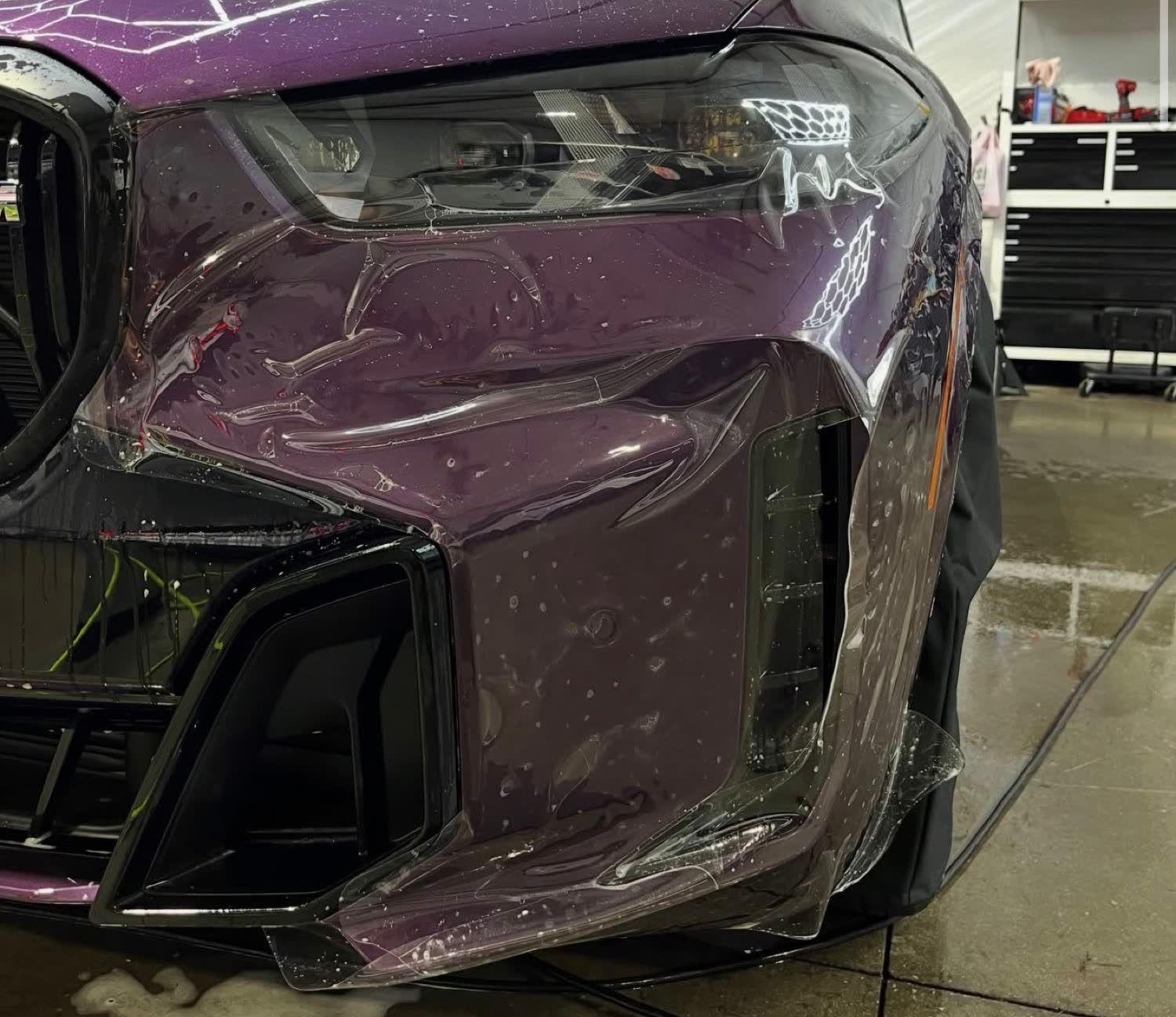 Front bumper of a purple BMW X5 with oversized paint protection film hanging past the edge before being wrapped and tucked
