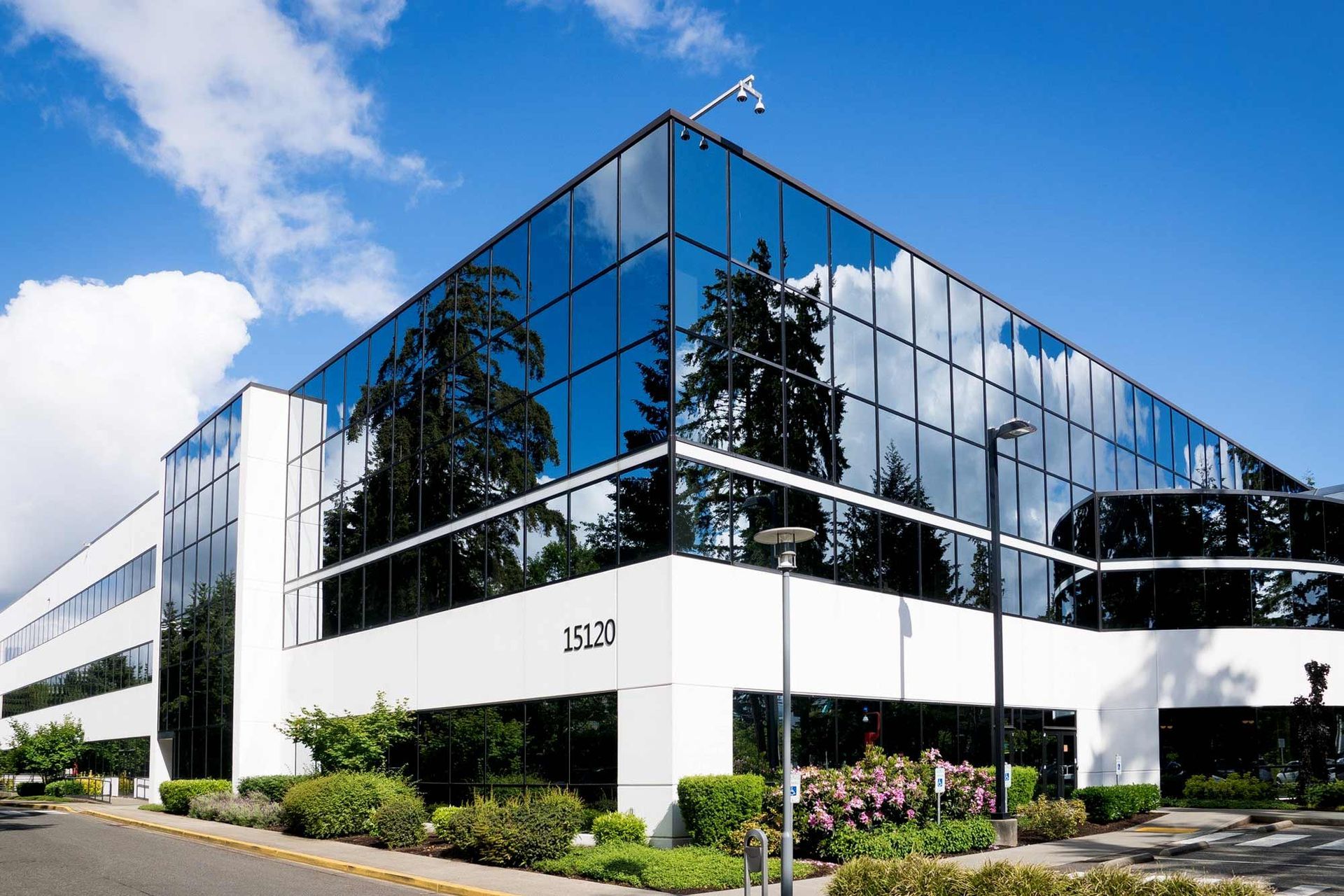 A three-story modern office building with a white base and a reflective blue-tinted glass facade under a bright sky.