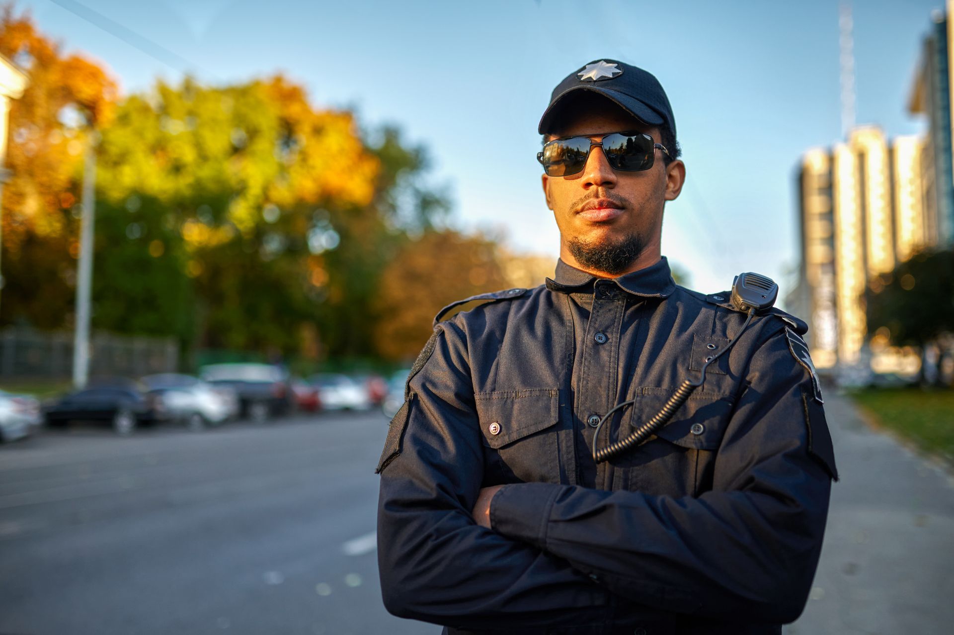 A security guard wearing sunglasses and a hat is standing on a sidewalk with his arms crossed.