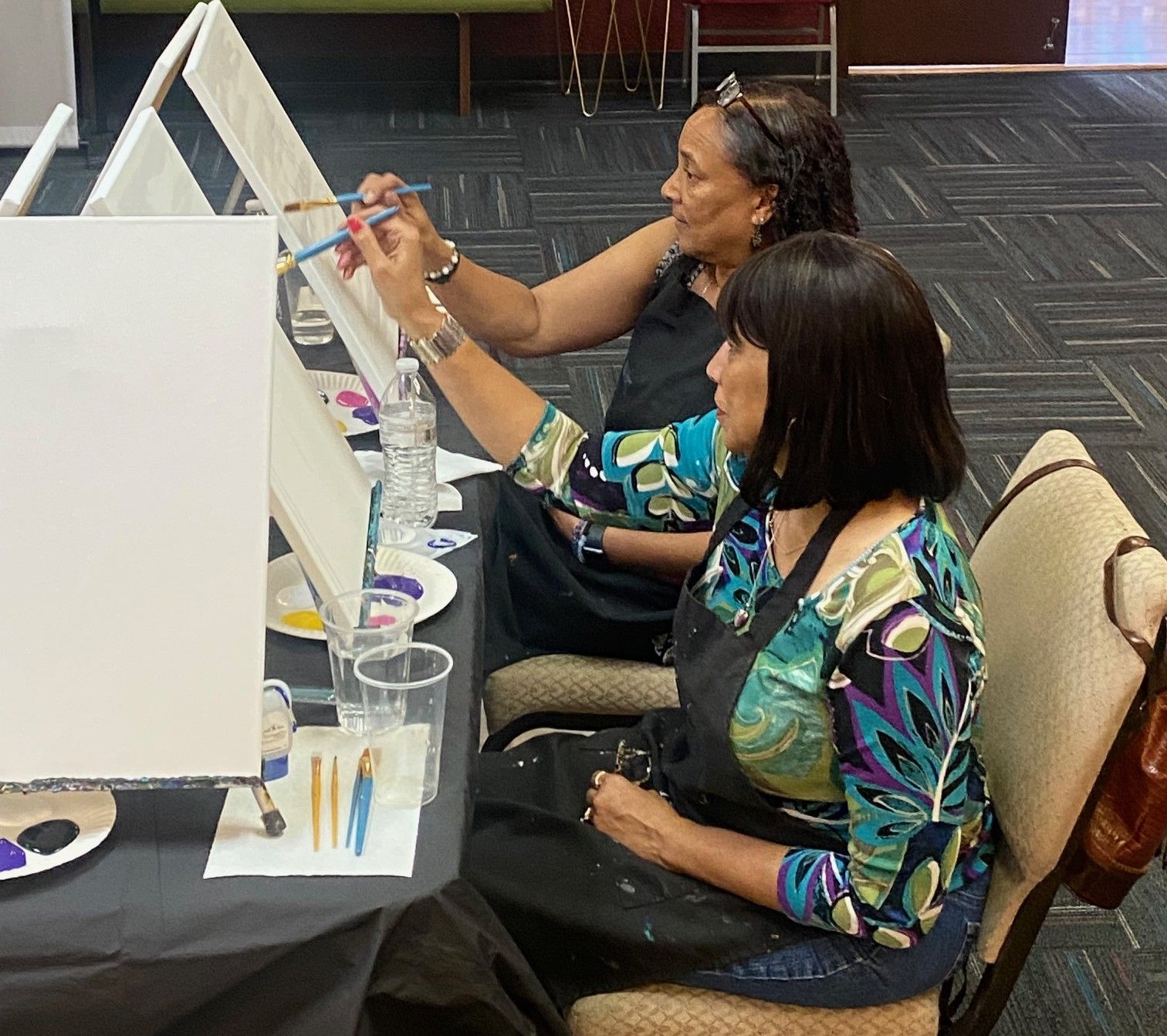 Two people wearing aprons sit at a table painting on canvases in an indoor workshop setting.
