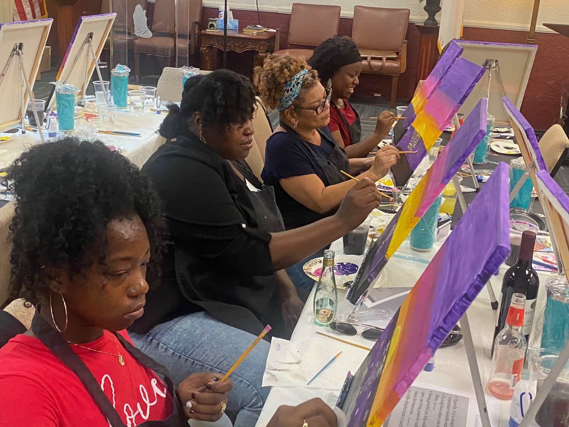 Four people sit at tables painting on canvases in a workshop, focusing on their colorful sunset-themed artwork.