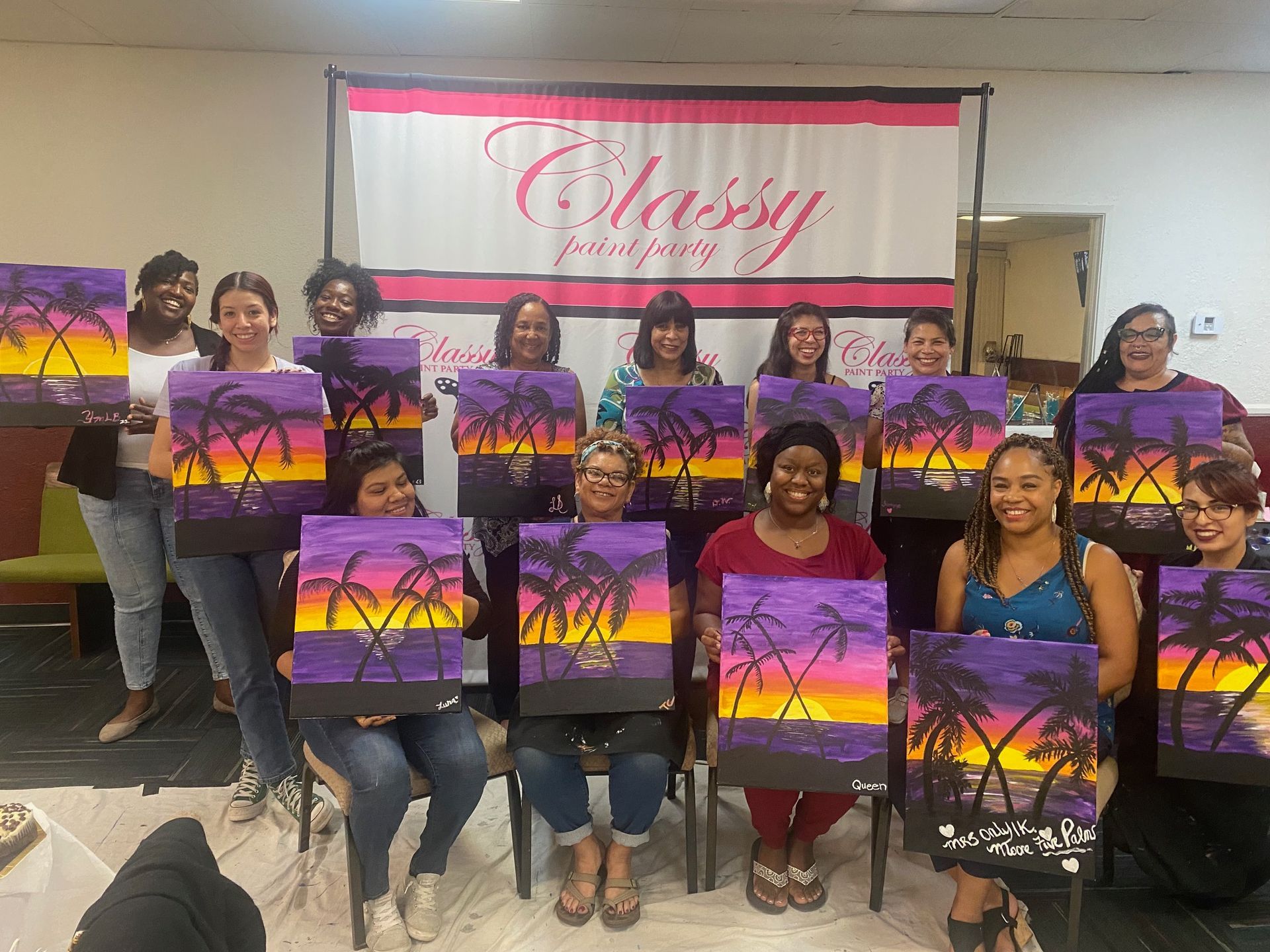A group of people holding their sunset palm tree paintings in front of a Classy Paint Party banner at a studio.