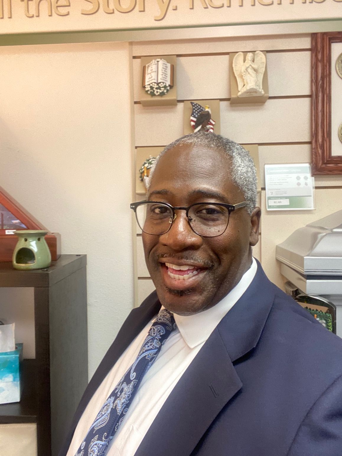 A smiling person in a blue suit, white shirt, and patterned blue tie standing in a room with wall decor.