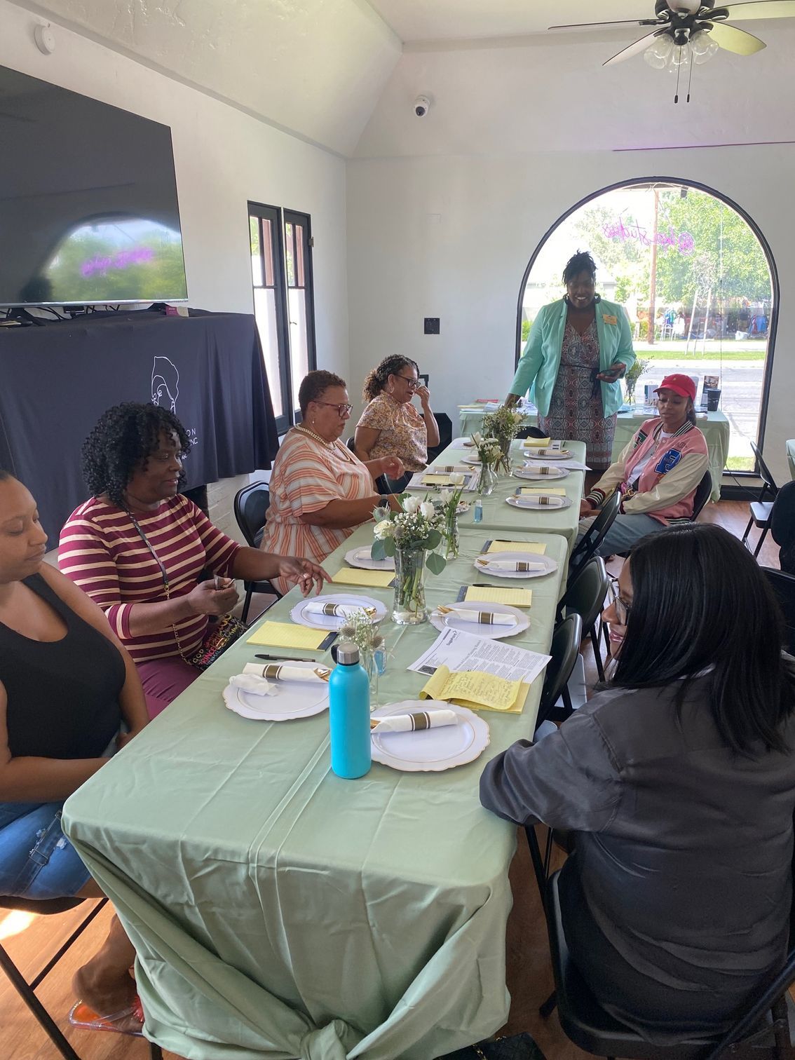 A group of people sit around a long dining table covered with a light green tablecloth for a meeting or gathering.