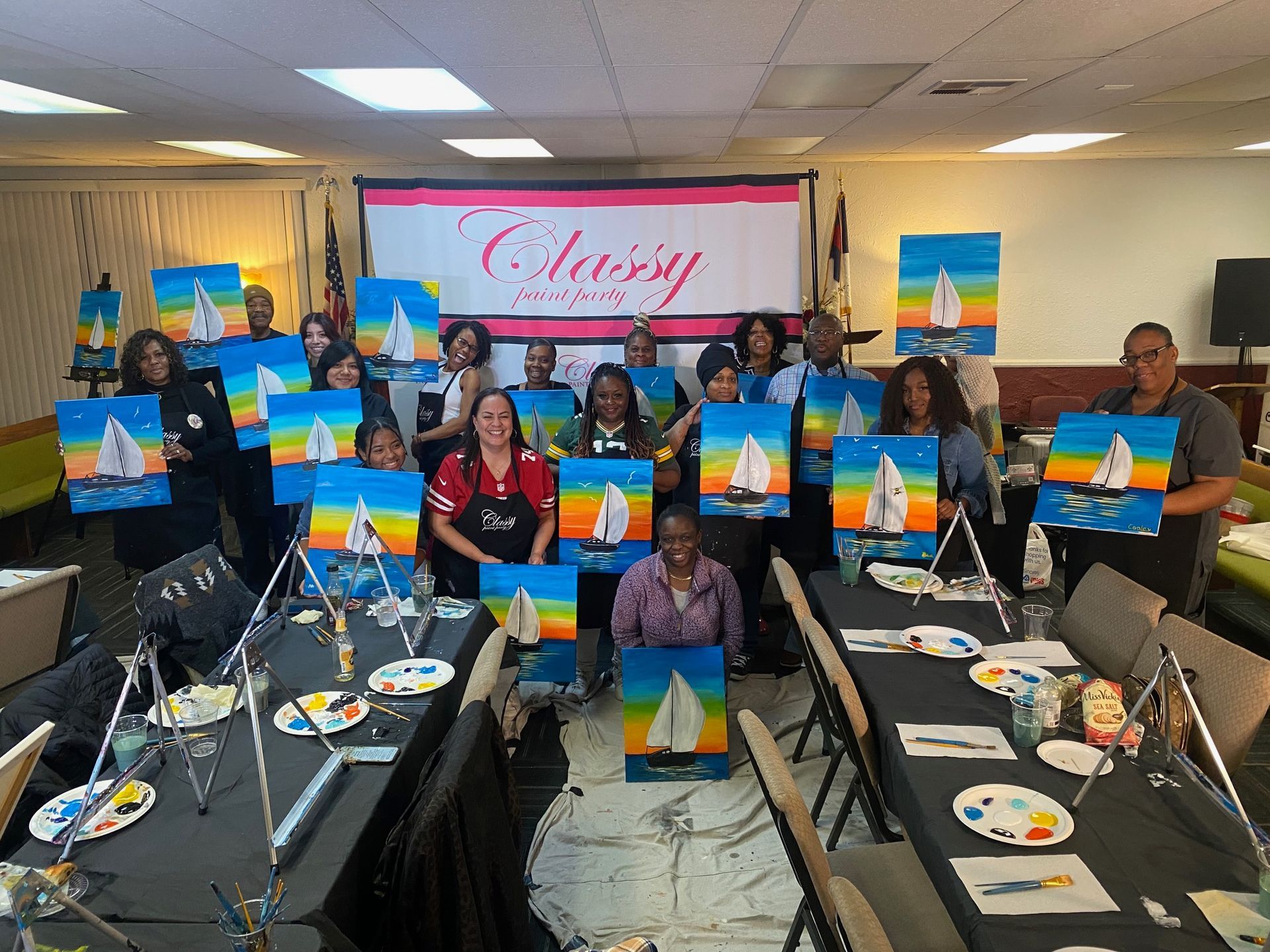 A group of people standing behind their easels, displaying sailboat paintings in a classroom with a 