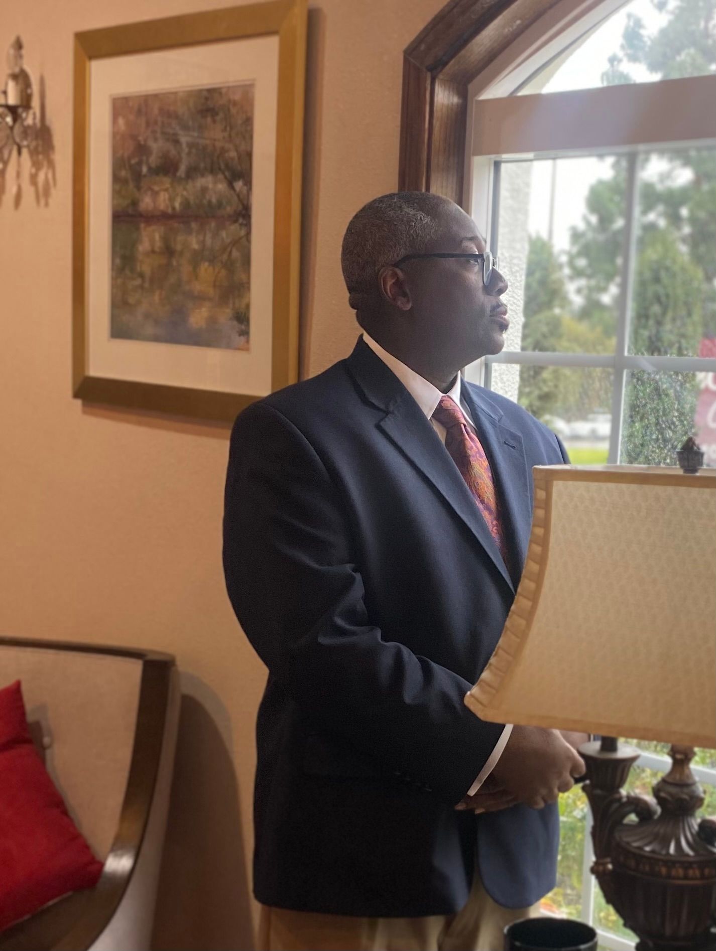 A person in a suit and tie stands by a window in a softly lit room, looking outside.