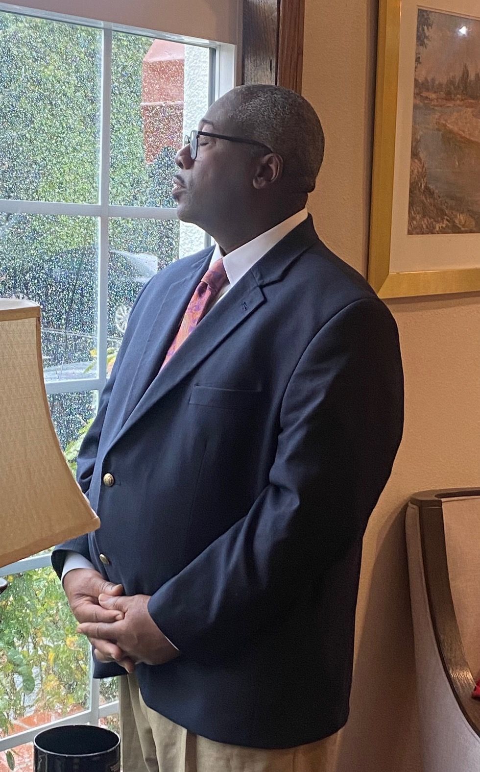 A person in a dark suit and red tie stands by a window, looking outside with their hands clasped.