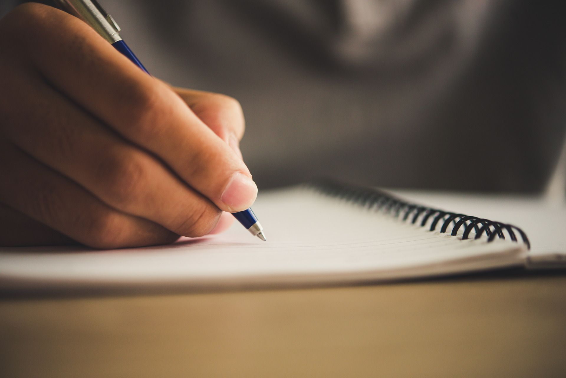 A person’s hand holds a blue pen and writes on the blank page of a spiral-bound notebook.