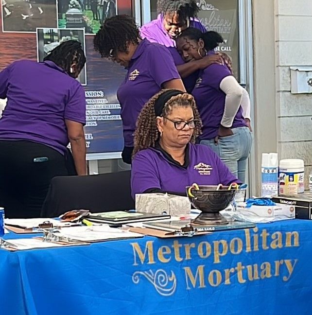 Four people wearing purple Metropolitan Mortuary shirts stand or sit behind a blue table at an outdoor event.