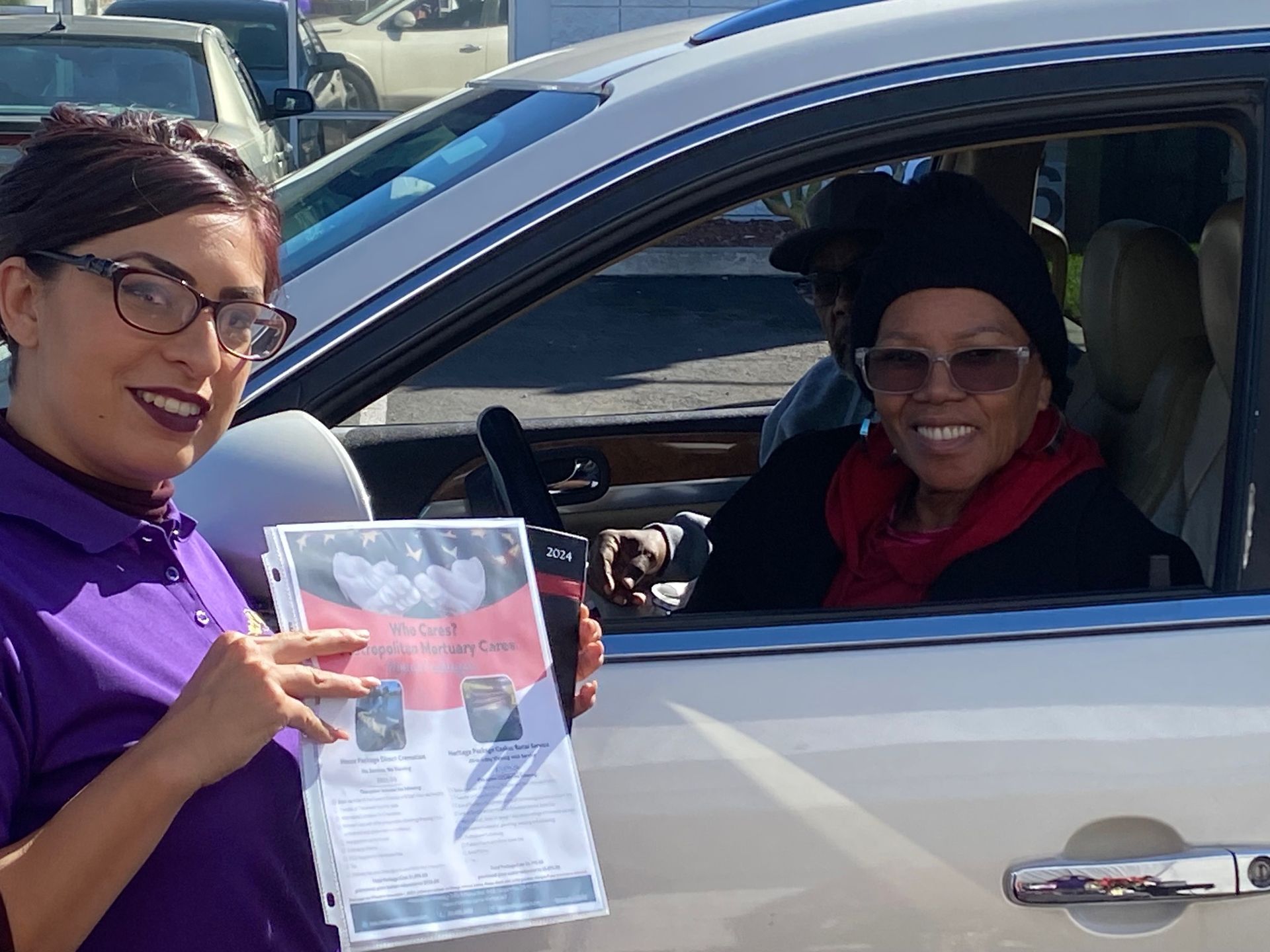A person in a purple shirt holds a document near a car window where an individual is seated and smiling.