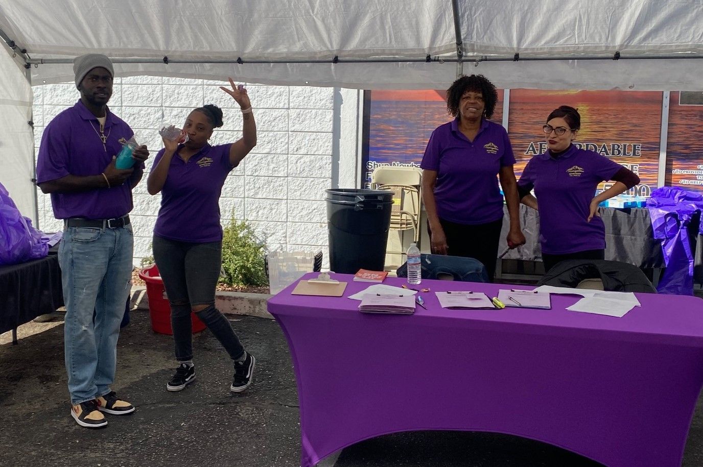 Four people in purple shirts stand behind a purple table at an outdoor event under a white tent.