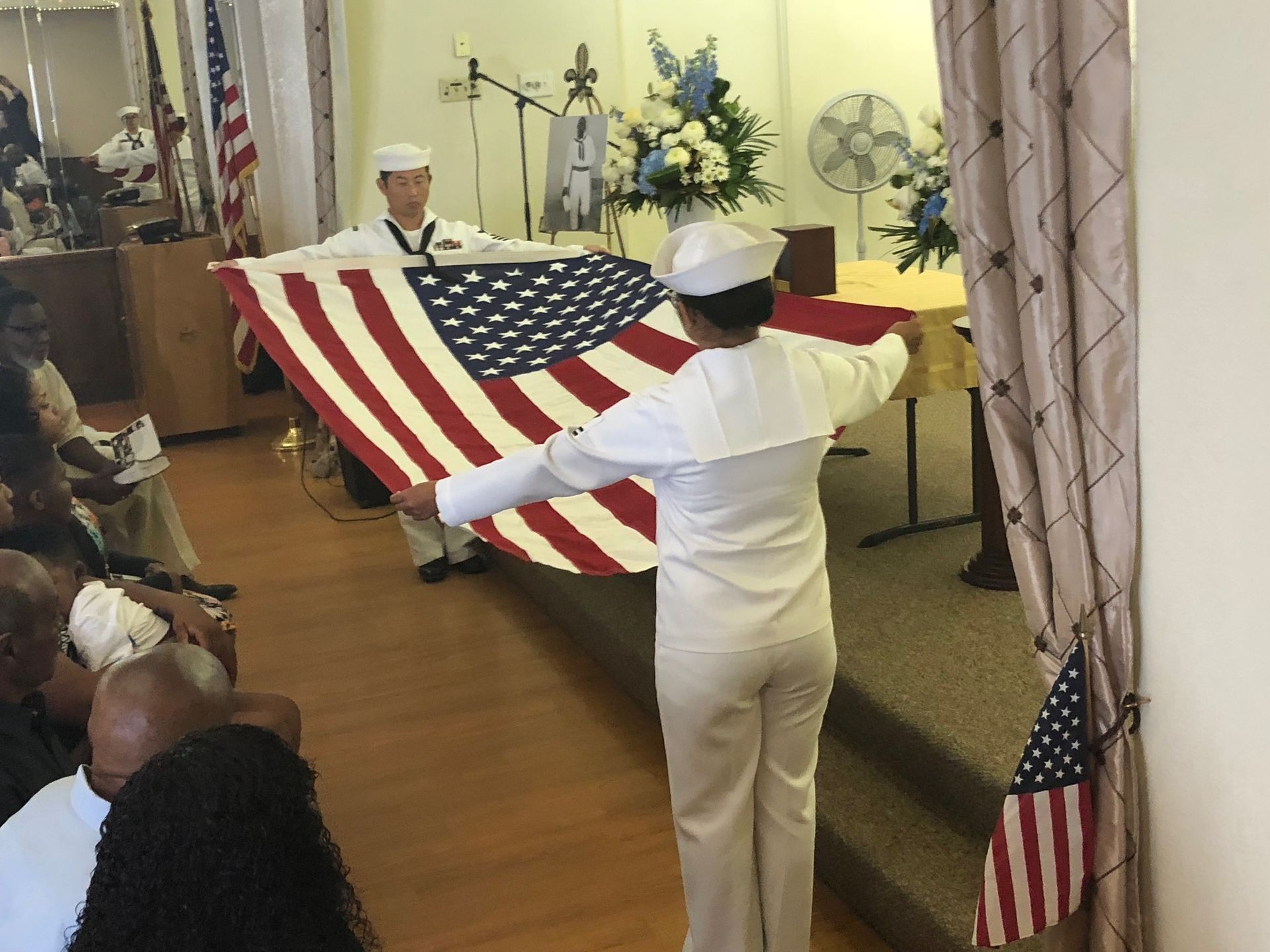 Two Navy service members in white uniforms hold an American flag during a ceremony in a room with guests.