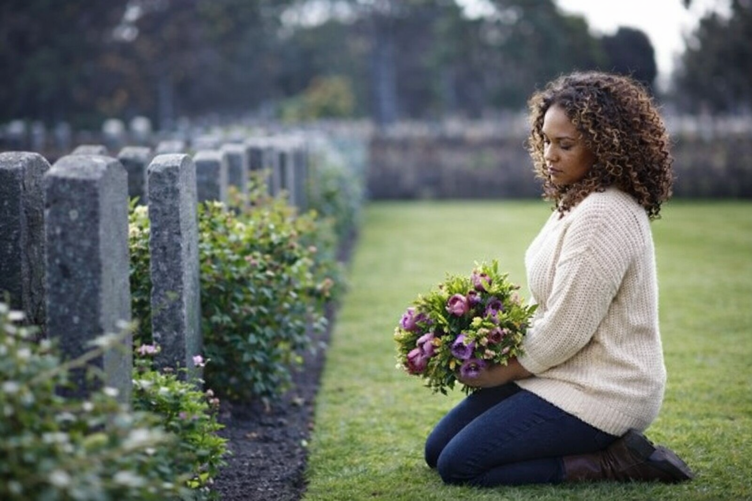 A person kneeling on the grass in a cemetery, holding a bouquet of purple flowers beside a row of stone markers.