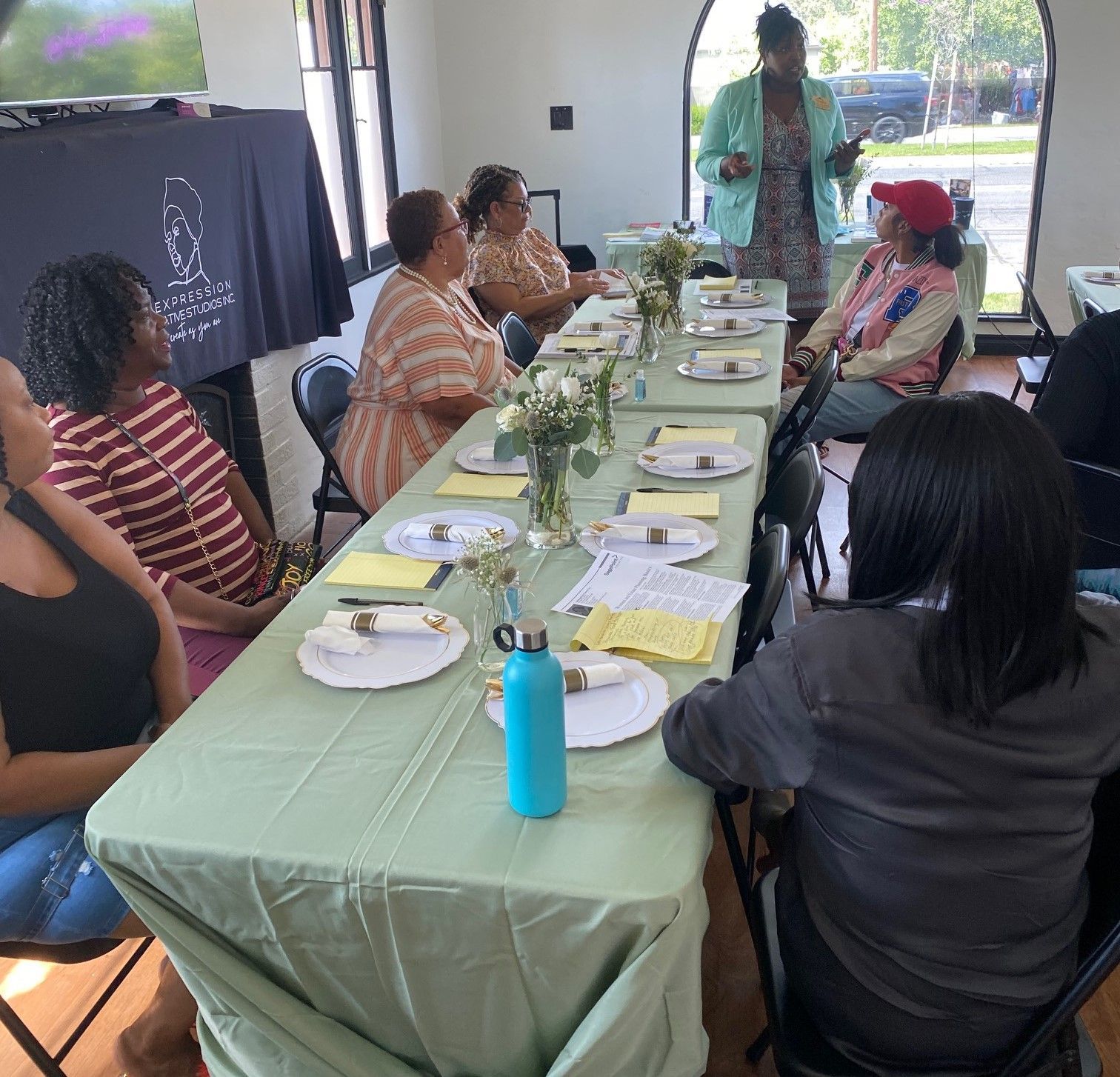 People sit around a long, light green-covered table in a room while a woman stands at the head of the table speaking.