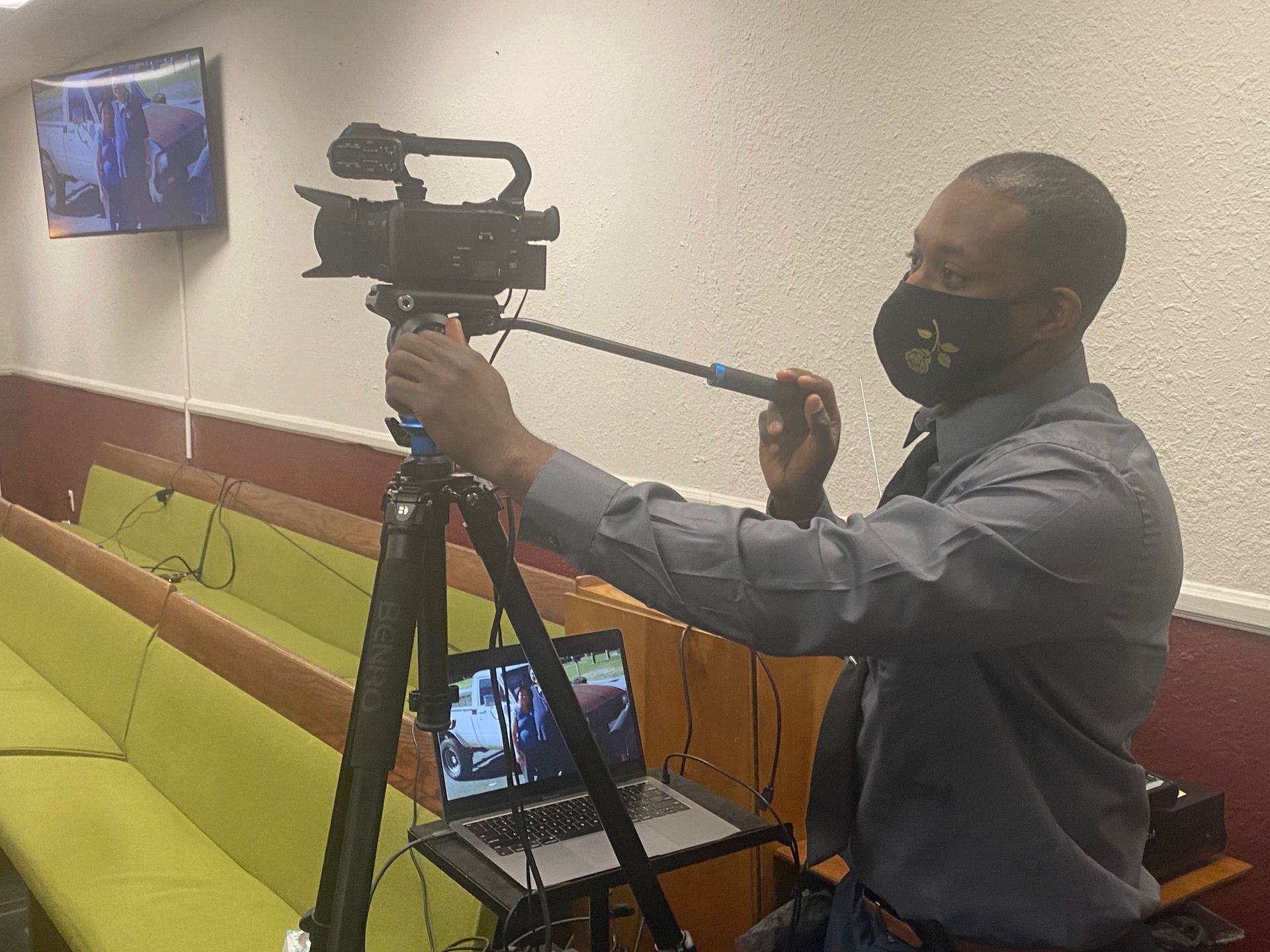 A person wearing a face mask operates a professional video camera on a tripod, with a laptop open on a stand nearby.