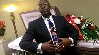 A person in a suit and patterned tie stands in front of an open casket decorated with flowers.