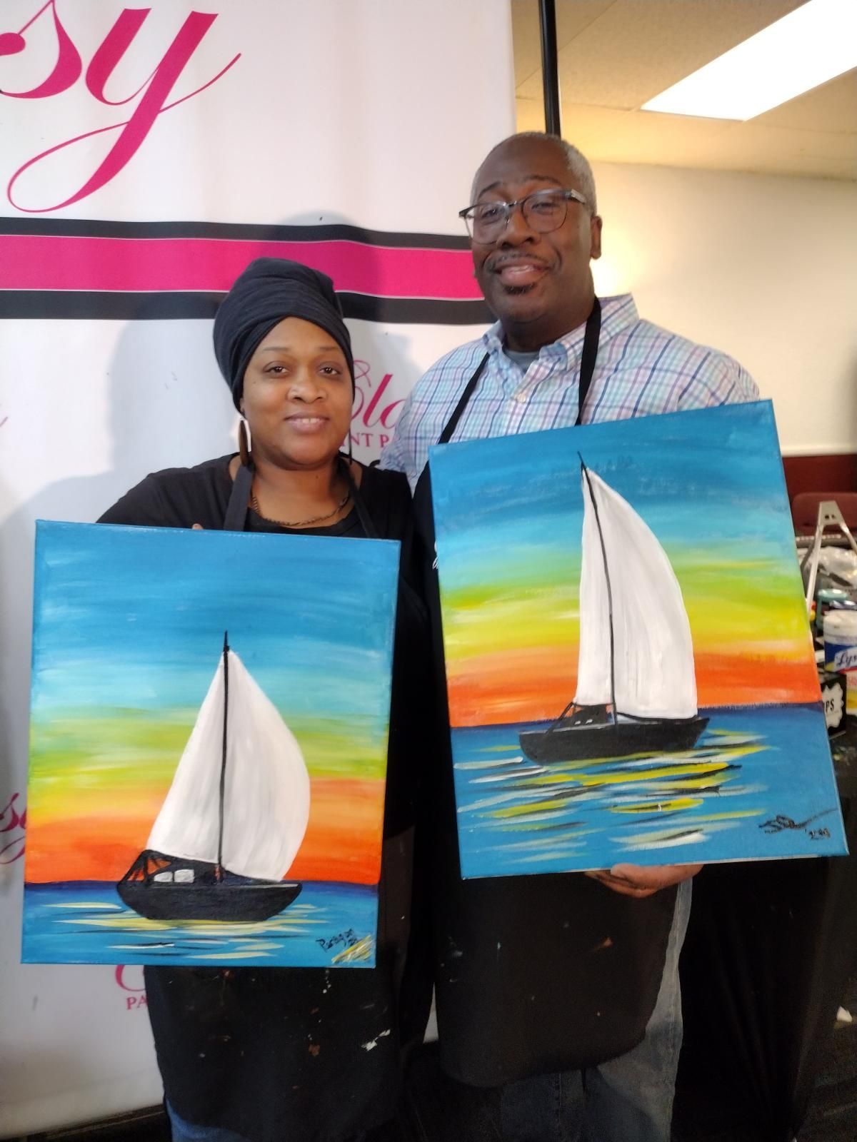 Two people smile while holding up their matching paintings of sailboats on a vibrant sunset sea at a painting event.