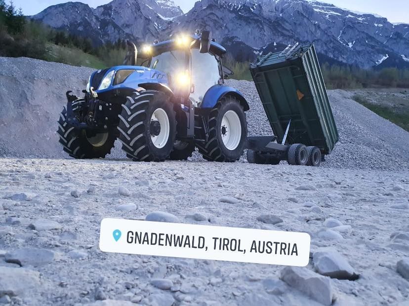 ein blauer traktor fährt auf einem felsigen feld mit einem grünen anhänger .