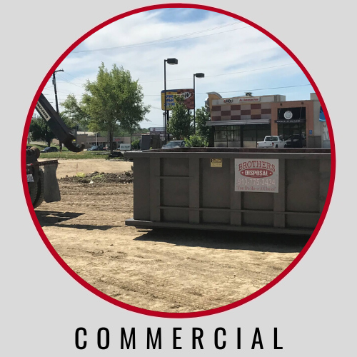 A large metal commercial dumpster sits on a dirt construction site in front of a small retail building.