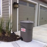 A grey wheeled trash bin with a red and white logo sits on a concrete driveway in front of a house with garage doors.