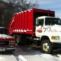 A white garbage truck with a bright red hopper parked on a snowy street, beside a red GMC pickup truck.