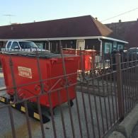 Two red industrial dumpsters sit in a parking lot behind a decorative metal fence, next to a building and a pickup truck.