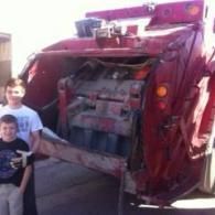 Two children standing next to the rear loading compartment of a large red garbage truck.