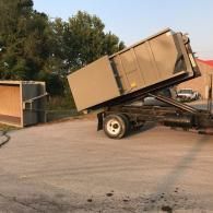A tan dumpster being loaded onto a flatbed trailer in a paved outdoor area.