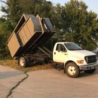 A white roll-off dump truck with its container raised, parked on a paved lot near trees.