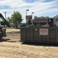 A large, gray industrial dumpster sits on a dirt lot near a commercial building with trees and light poles in the distance.