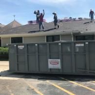Three workers repair a building roof, standing near a large, grey dumpster in a parking lot under a bright blue sky.