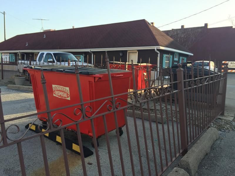 A red metal dumpster sits behind a brown metal fence in front of a small, single-story commercial building.