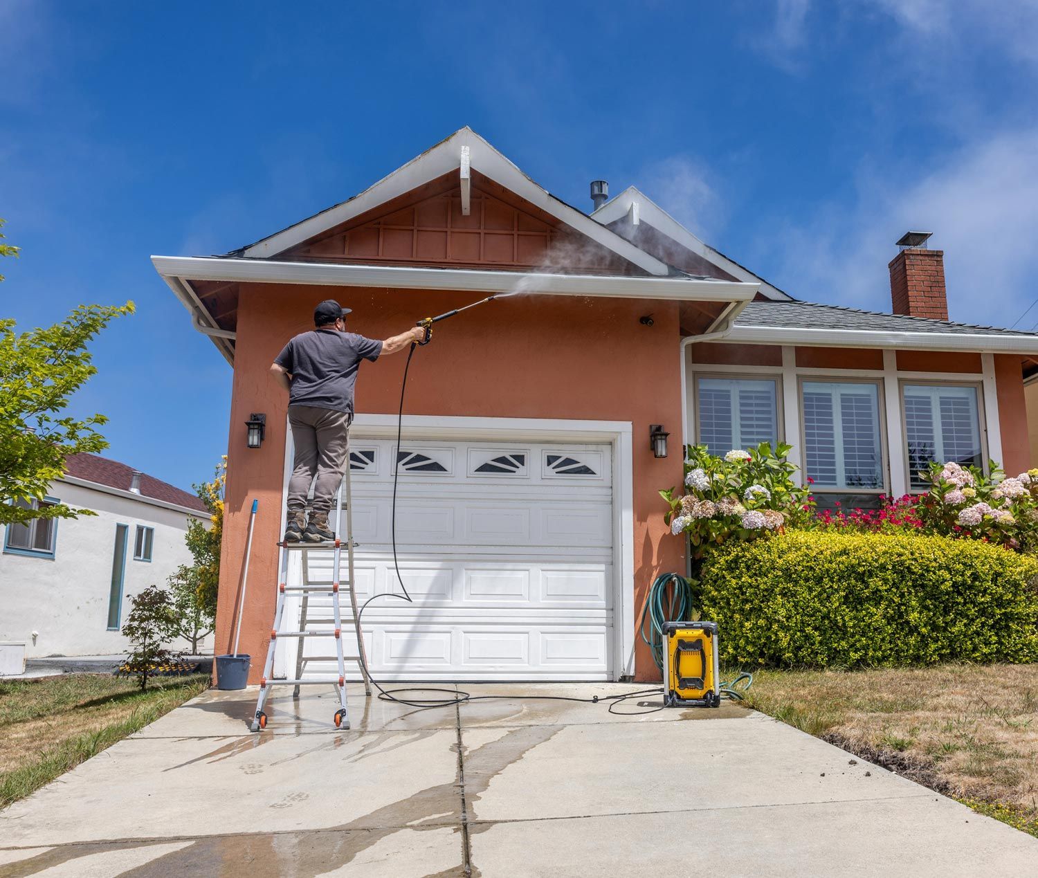 A man is standing on a ladder cleaning a house with a high pressure washer.
